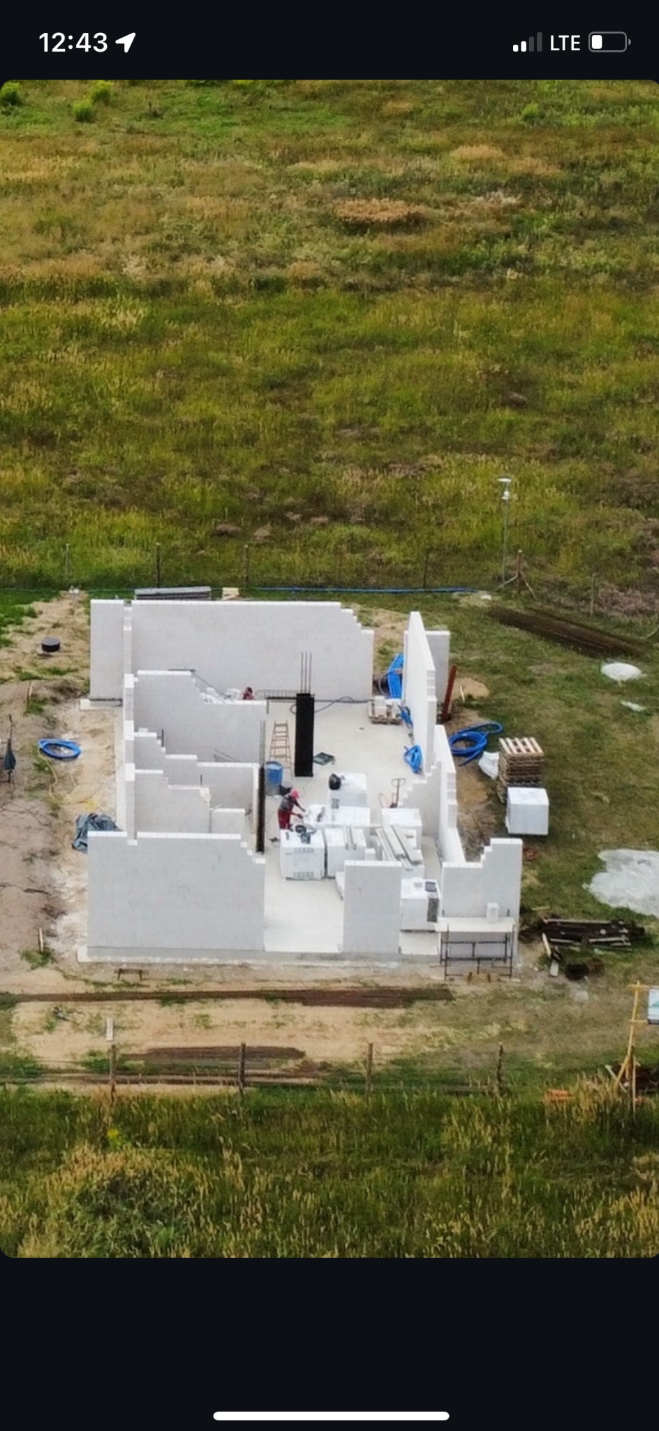 Budowa domu z białych bloczków, widok z góry na częściowo wzniesione ściany. Na budowie pracownicy w czerwonych ubraniach i sprzęt budowlany. Trawa w tle.