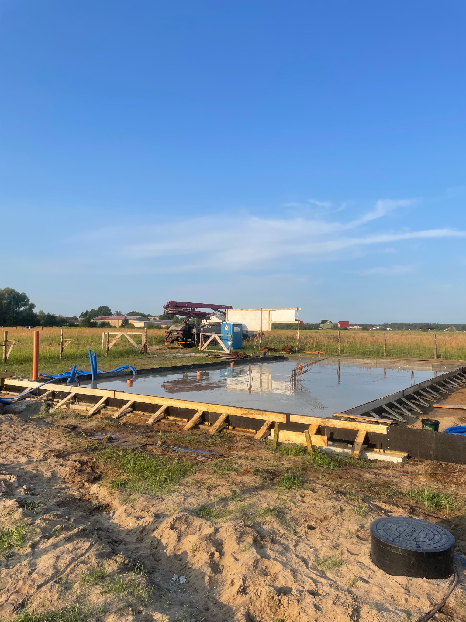 Wylany fundament pod budowę domu jednorodzinnego. Widoczna drewniana szalówka, zbrojenie i świeży beton. W tle maszyna do betonu i zielone pola.