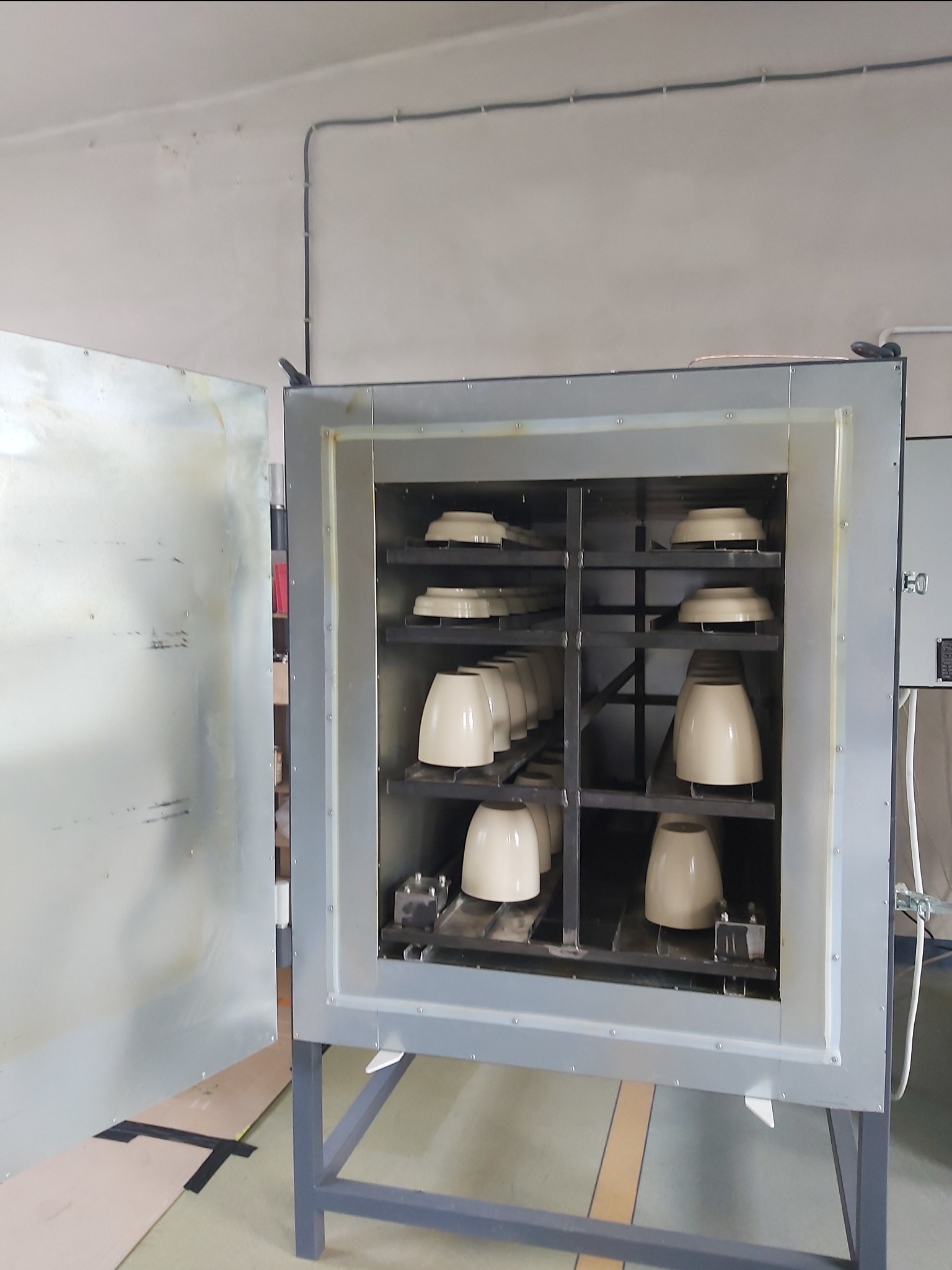 Metalowa komora pieca z otwartymi drzwiami, wewnątrz ułożone na półkach ceramiczne elementy w kształcie kloszy i pierścieni, przygotowane do procesu obróbki termicznej.