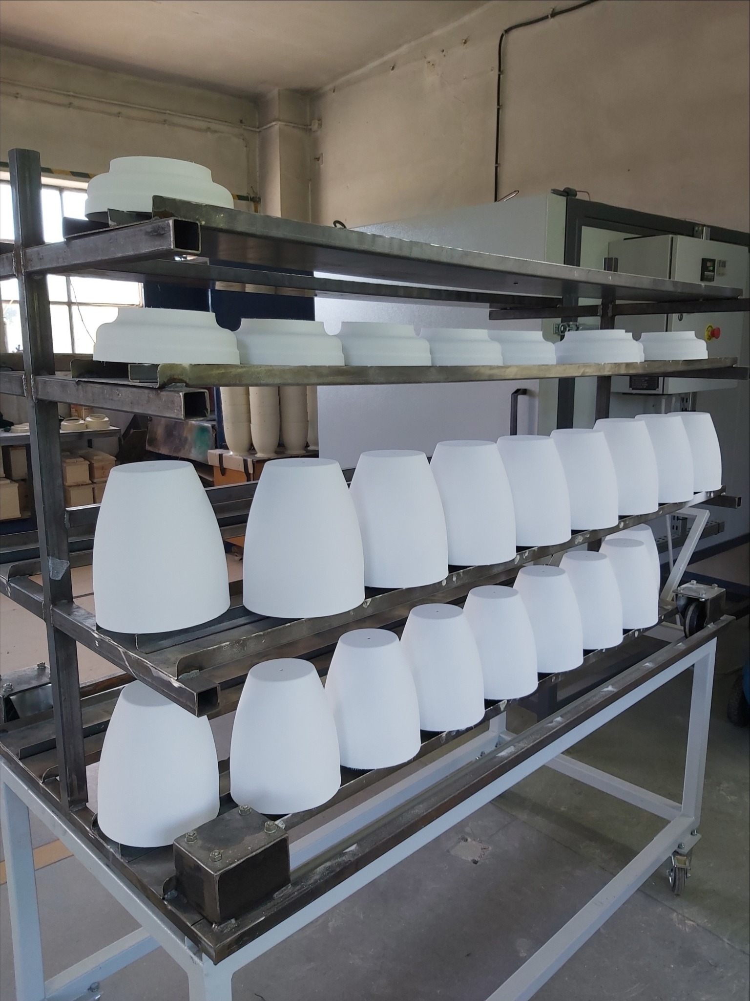 Metalowy regał na kółkach z białymi, stożkowymi elementami oświetleniowymi po procesie malowania, widoczny piec do wygrzewania w tle.