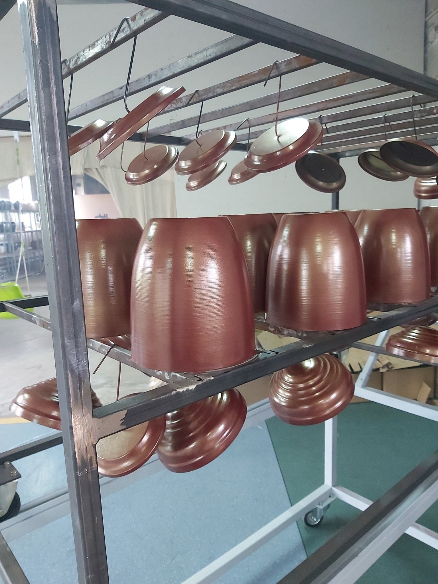 Metalowe elementy, prawdopodobnie donice i podstawki, po procesie malowania proszkowego w odcieniach miedzi, wiszące na metalowym stelażu w hali produkcyjnej.