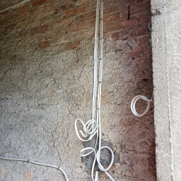 GREMONT Grzegorz Burski - Białe przewody elektryczne prowadzone po surowej, częściowo otynkowanej ścianie z cegły, zabezpieczone uchwytami, z widocznym fragmentem surowego otworu w murze.