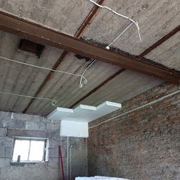 GREMONT Grzegorz Burski - Wnętrze w trakcie prac remontowych: widoczny surowy betonowy sufit z metalowymi belkami, nowa instalacja elektryczna w białych peszlach oraz częściowo zamontowane płyty styropianowe do izolacji...