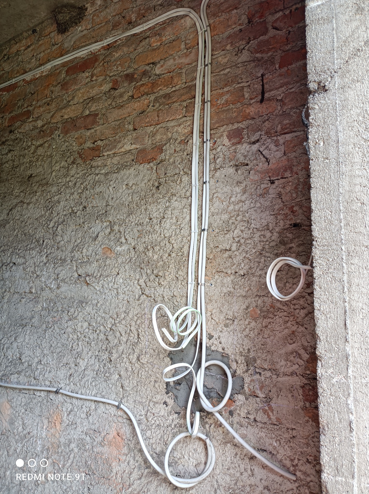 Białe przewody elektryczne prowadzone po surowej, częściowo otynkowanej ścianie z cegły, zabezpieczone uchwytami, z widocznym fragmentem surowego otworu w murze.