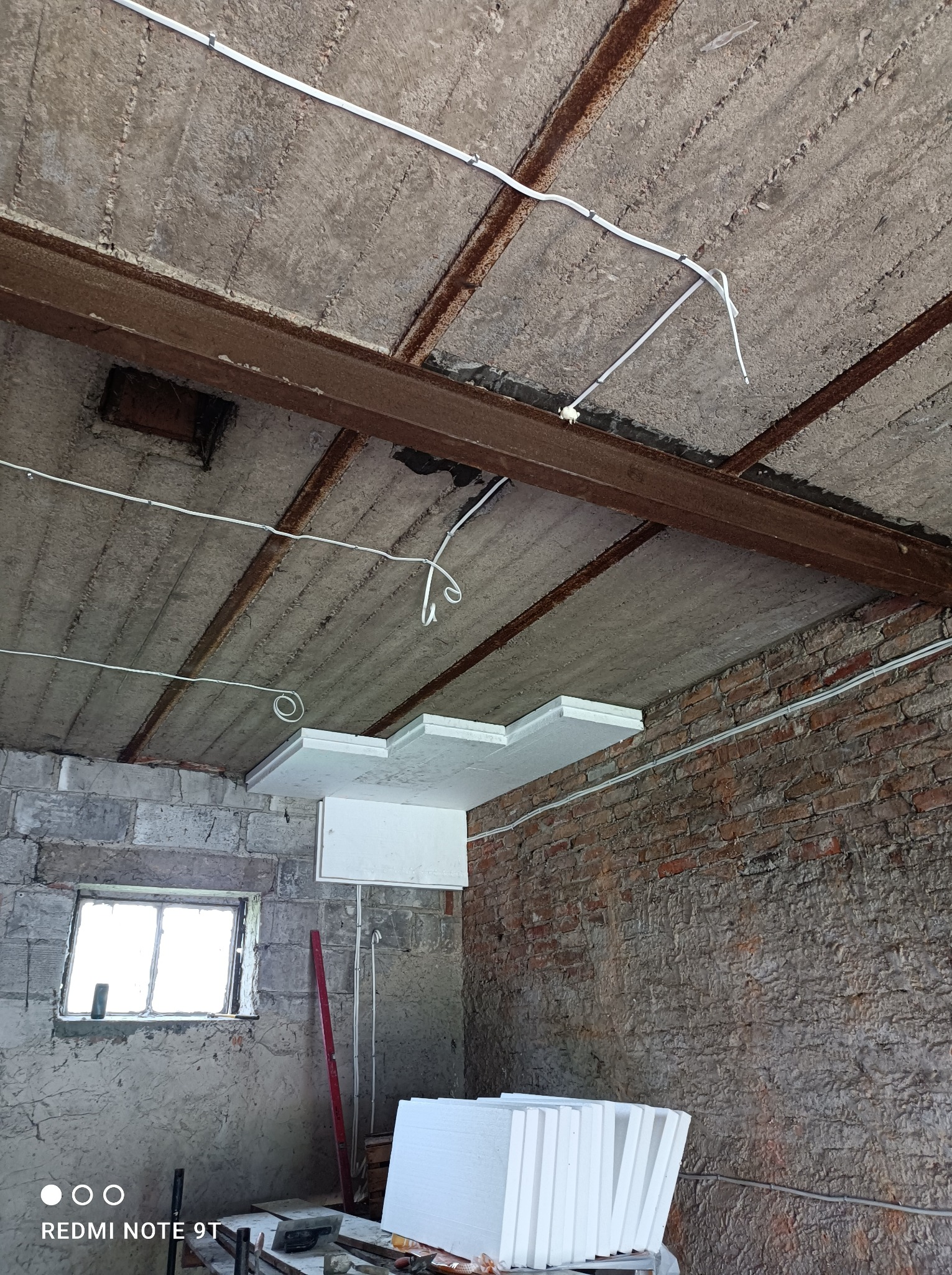 Wnętrze w trakcie prac remontowych: widoczny surowy betonowy sufit z metalowymi belkami, nowa instalacja elektryczna w białych peszlach oraz częściowo zamontowane płyty styropianowe do izolacji...