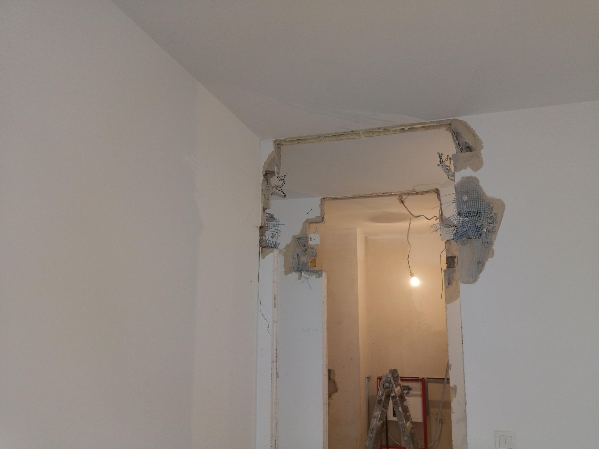 Otwór drzwiowy w trakcie remontu, widoczne fragmenty usuniętego muru, siatka wzmacniająca i wystające przewody elektryczne, w tle drabina i żarówka.