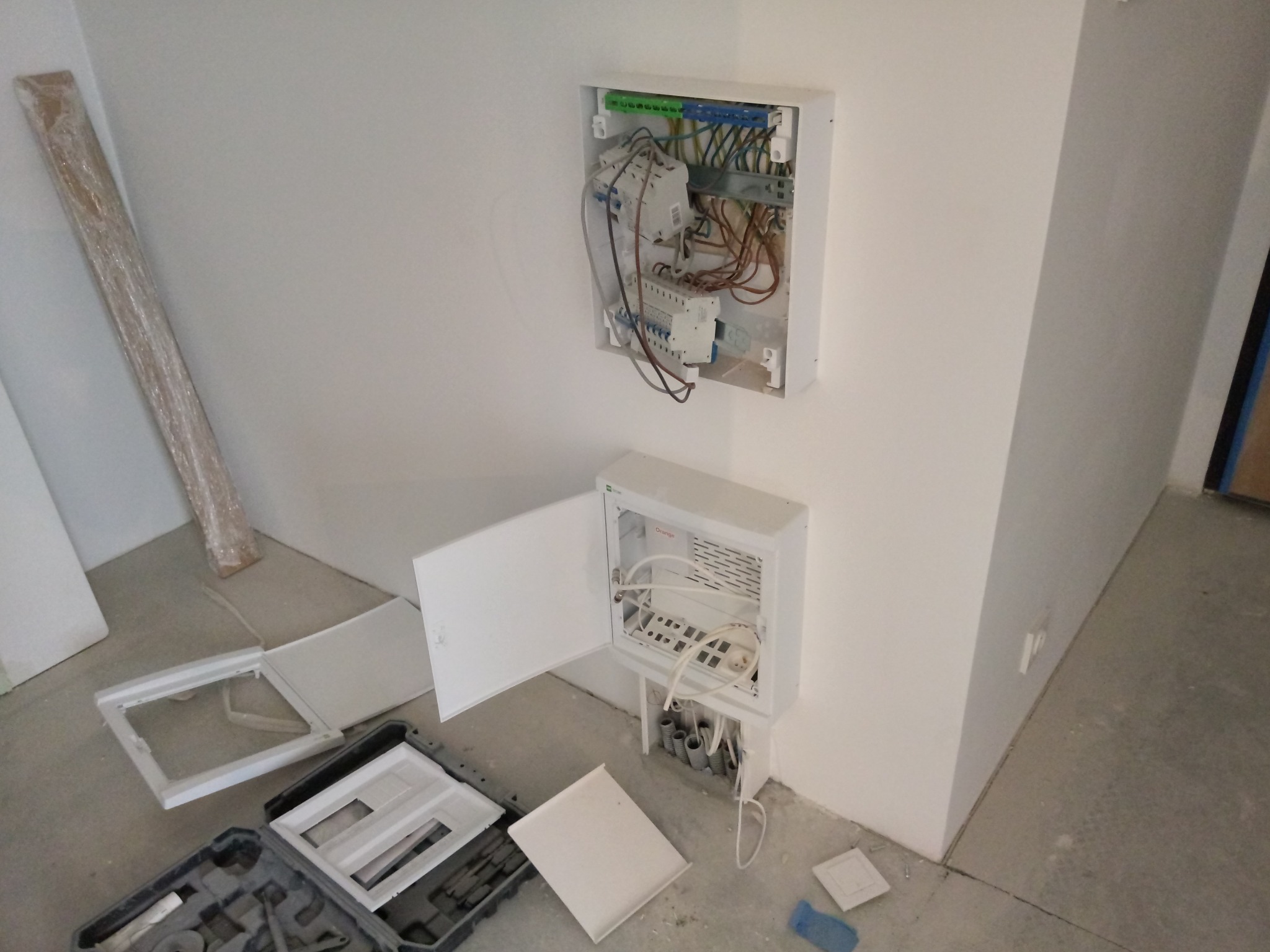 Rozdzielnica elektryczna w trakcie montażu na ścianie, z otwartą obudową, widoczne kable i przewody, skrzynka narzędziowa na podłodze, etap prac instalacyjnych w surowym wnętrzu.