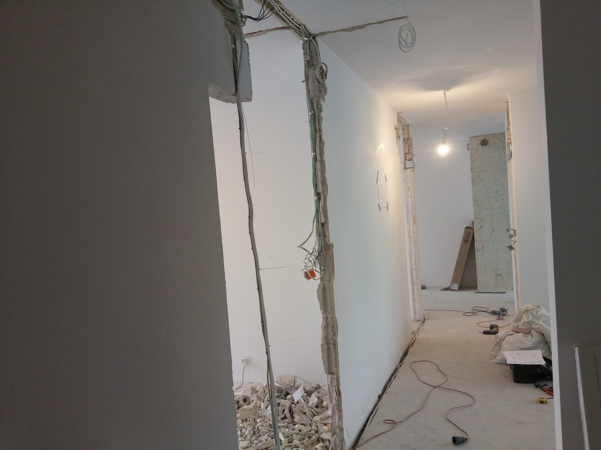 Wnętrze w trakcie remontu: odsłonięte przewody elektryczne w ścianie, gruz, narzędzia na podłodze, widok na korytarz z wiszącą żarówką.
