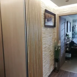 Wnętrze przedpokoju z dekoracyjną ścianą z fakturą kamienia, drewnianą zabudową z lamelami i przesuwnymi drzwiami oraz obrazem w ciemnej ramie.