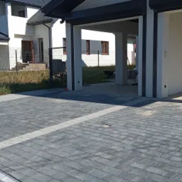 Szara, prostokątna nawierzchnia z kostki, z jasnym pasem wzdłuż, prowadząca do garażu w nowoczesnym domu z szarą blachodachówką i ogrodzeniem panelowym w tle.