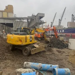 Żółta koparka kołowa Komatsu PW160 na błotnistym placu budowy, w tle pomarańczowa koparka, betonowe kręgi i niebieskie rury, widok na port z dźwigami i statkiem, pochmurne niebo.