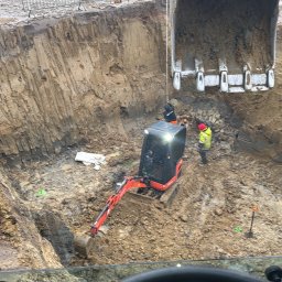 Usługi koparko-ladowarka Oliver Konkolewski - Widok z kabiny maszyny na wykop z pracującą minikoparką, operator w kasku, drugi pracownik w wykopie, geodeta na krawędzi wykopu, pochmurny dzień.