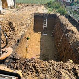 Usługi koparko-ladowarka Oliver Konkolewski - Głęboki wykop w ziemi, widoczna drabina, poziomica laserowa i narzędzia, w tle budynek w trakcie budowy i zielone pole, ujęcie z kabiny maszyny budowlanej.