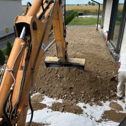 Usługi koparko-ladowarka Oliver Konkolewski - Pomarańczowa koparka CASE nabiera ziemię na białej geowłókninie, widoczny operator z łopatą, przygotowanie terenu pod budowę domu z dużym oknem.