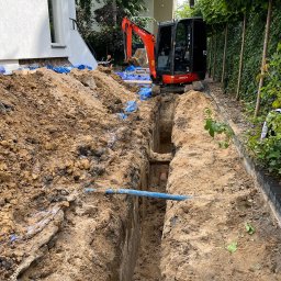 Usługi koparko-ladowarka Oliver Konkolewski - Wąski wykop w ziemi, z widoczną rurą wewnątrz i małą koparką Kubota obok, zabezpieczony niebieską folią, na tle budynku i zieleni.