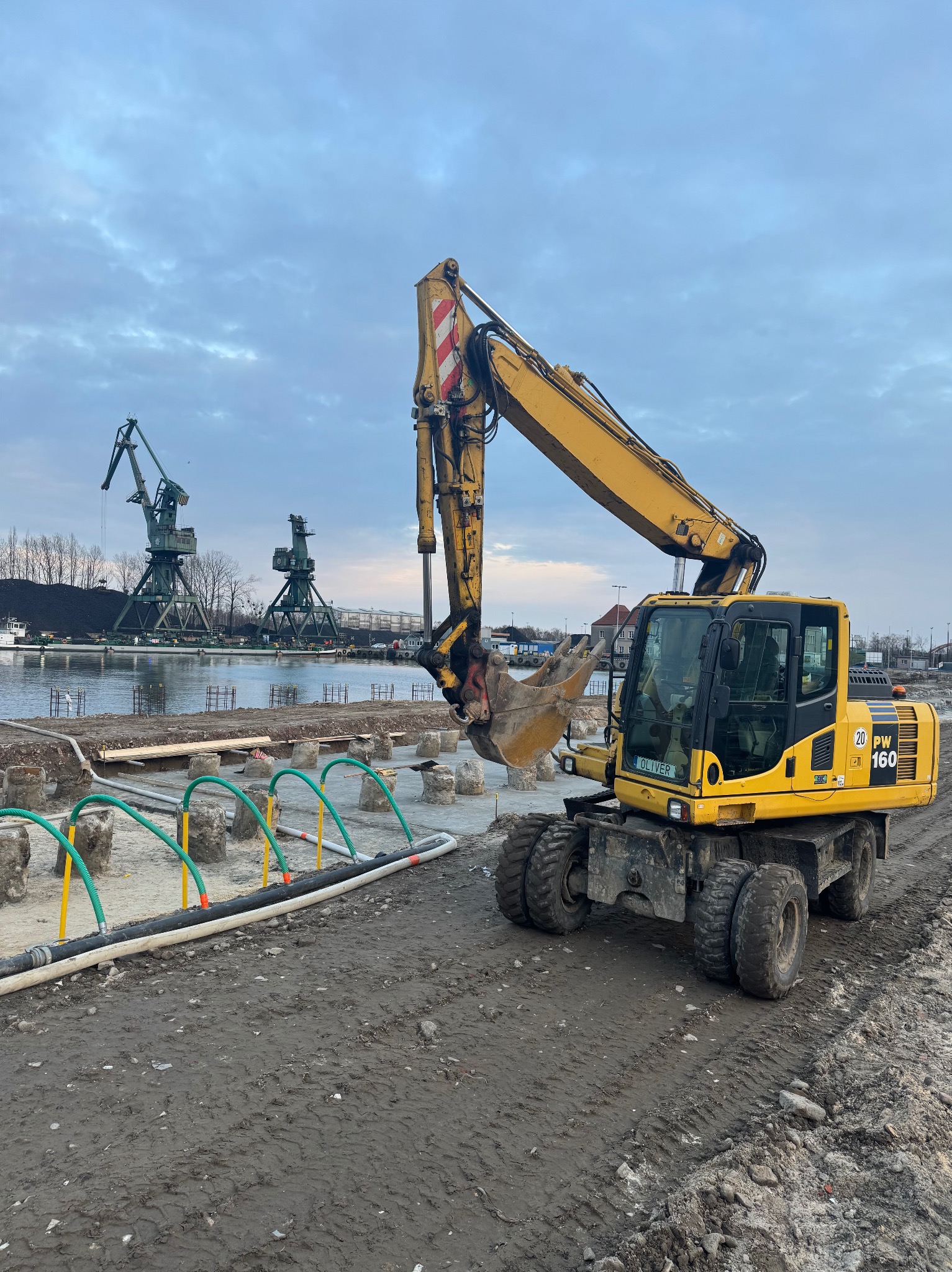 Żółta koparka kołowa Komatsu PW160 na placu budowy, w tle nabrzeże z dźwigami portowymi i zbiorowiskiem węgla, widoczne przygotowania do fundamentów.