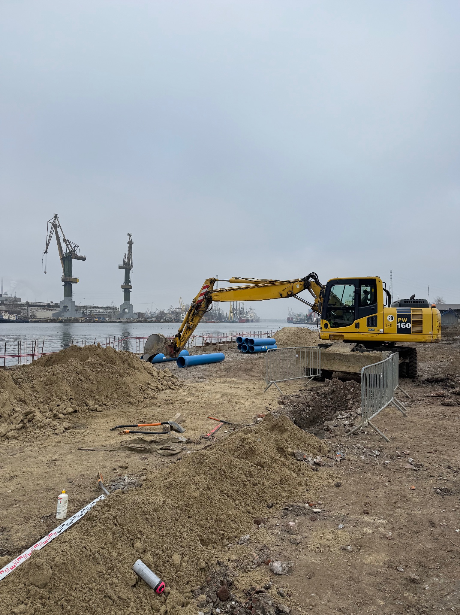 Żółta koparka Komatsu PW160 na placu budowy z rurami, w tle dźwigi portowe i pochmurne niebo.