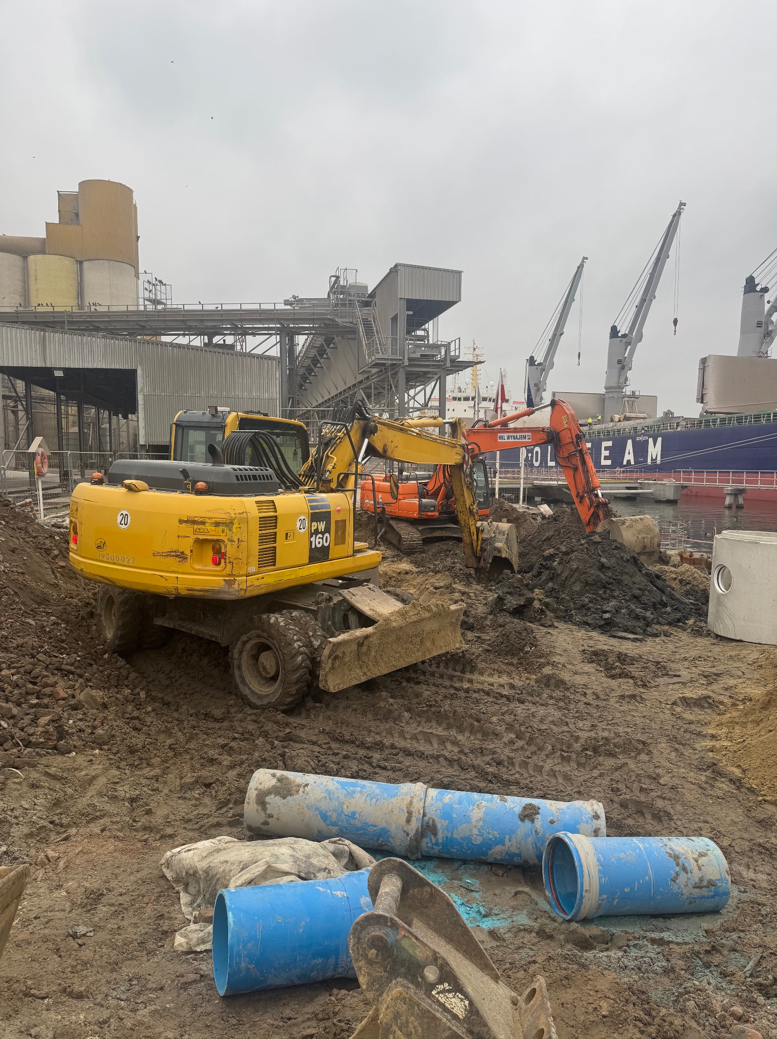 Żółta koparka kołowa Komatsu PW160 na błotnistym placu budowy, w tle pomarańczowa koparka, betonowe kręgi i niebieskie rury, widok na port z dźwigami i statkiem, pochmurne niebo.