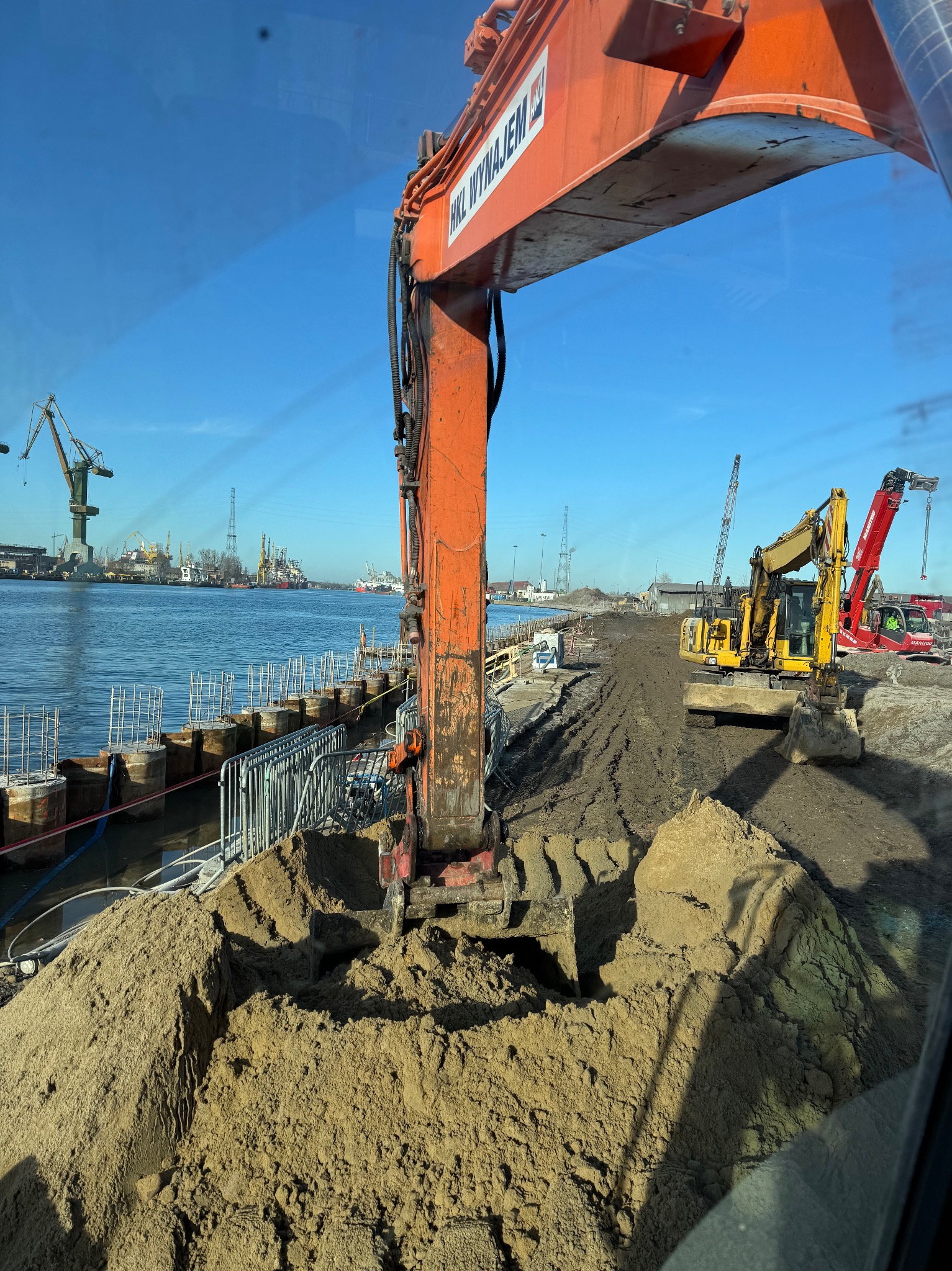 Pomarańczowa koparka zagłębia się w piasek na nabrzeżu, w tle widać inne maszyny budowlane, dźwigi portowe i zbiorniki wodne.