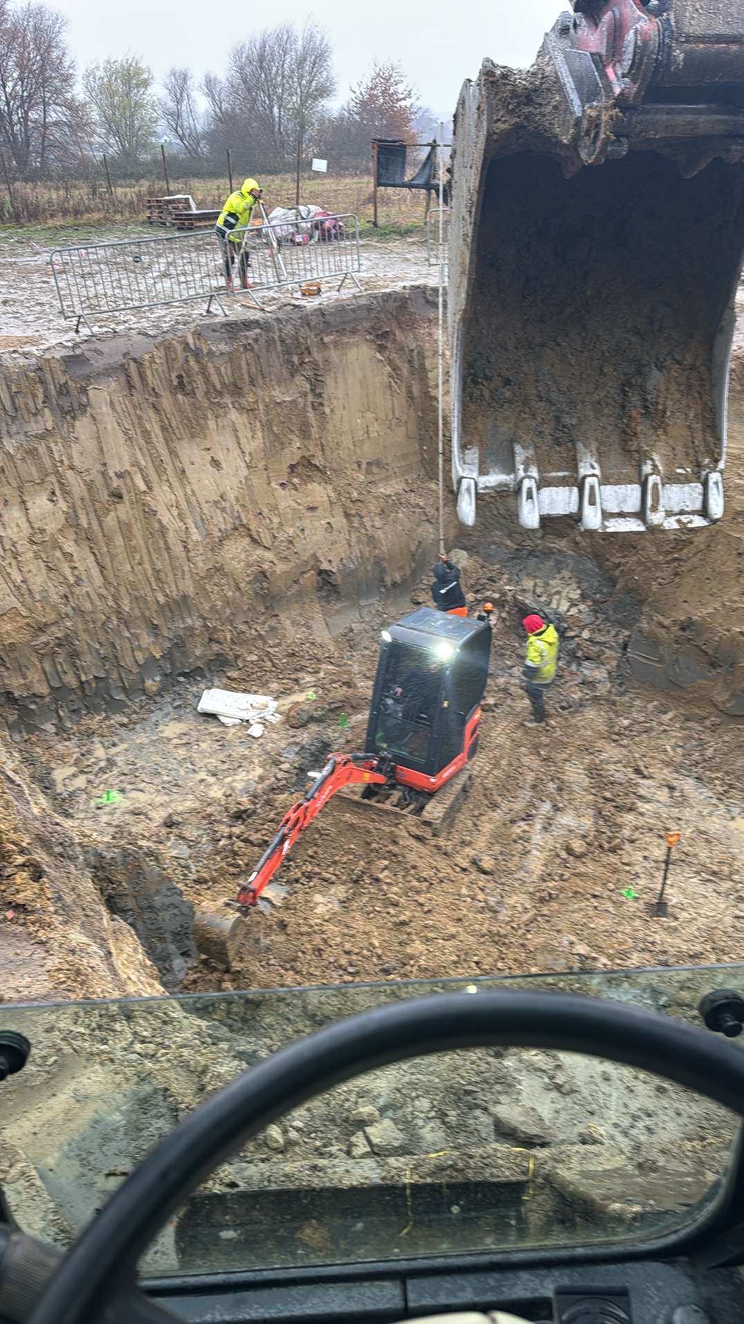 Widok z kabiny maszyny na wykop z pracującą minikoparką, operator w kasku, drugi pracownik w wykopie, geodeta na krawędzi wykopu, pochmurny dzień.