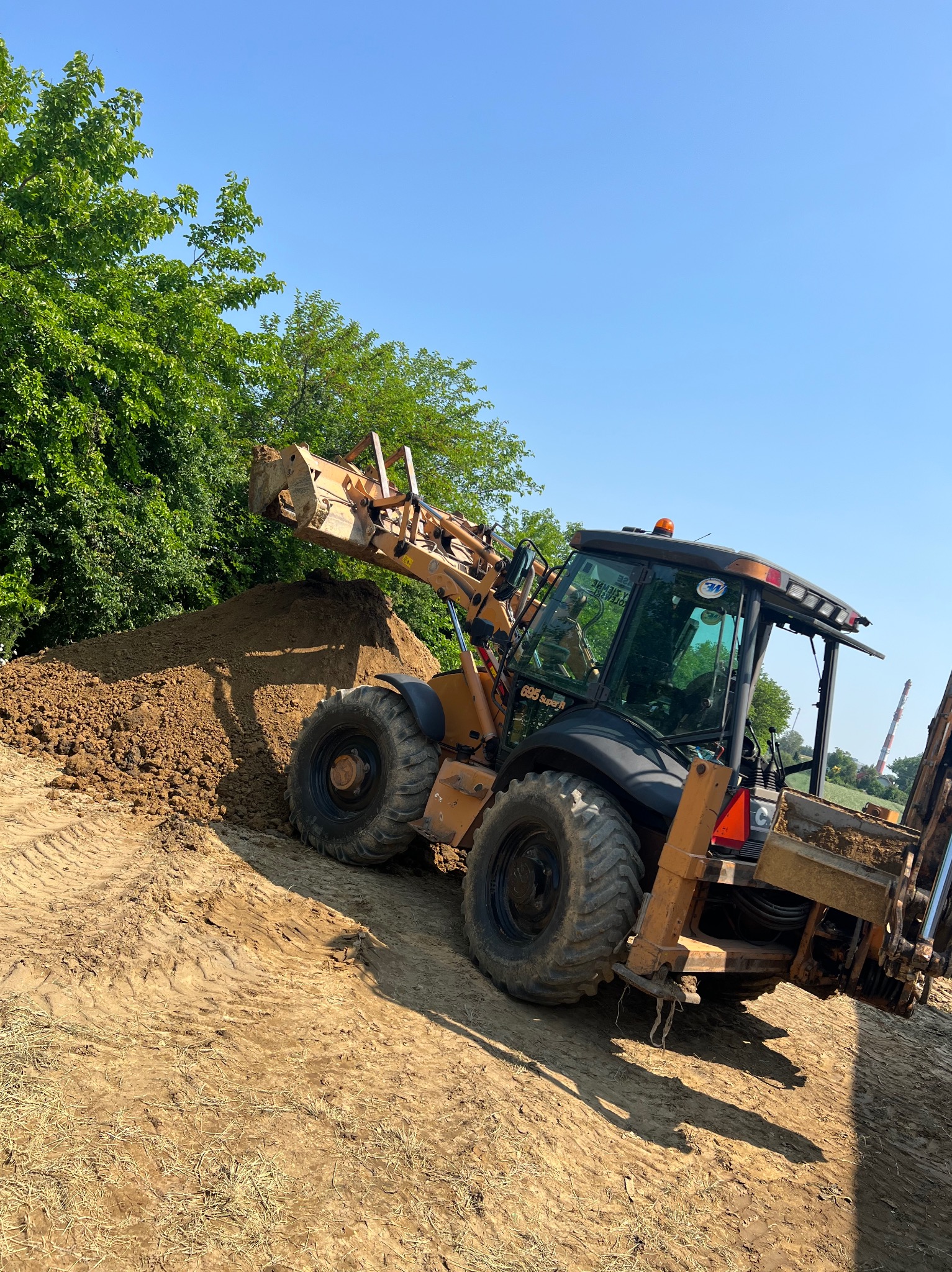 Żółto-czarna koparko-ładowarka Case 695 Super R z uniesioną łyżką nabierającą ziemię z pryzmy na placu budowy, w tle zielone drzewa i błękitne niebo.