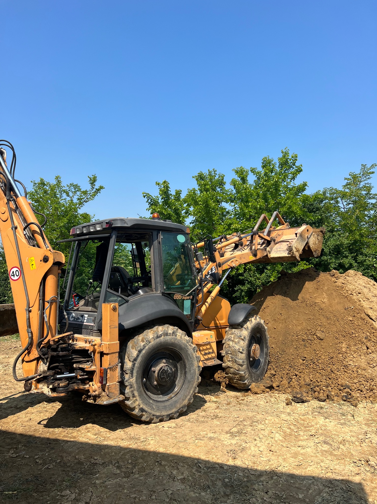 Pomarańczowa koparko-ładowarka marki Case nabiera ziemię z pryzmy na tle zieleni i błękitnego nieba.