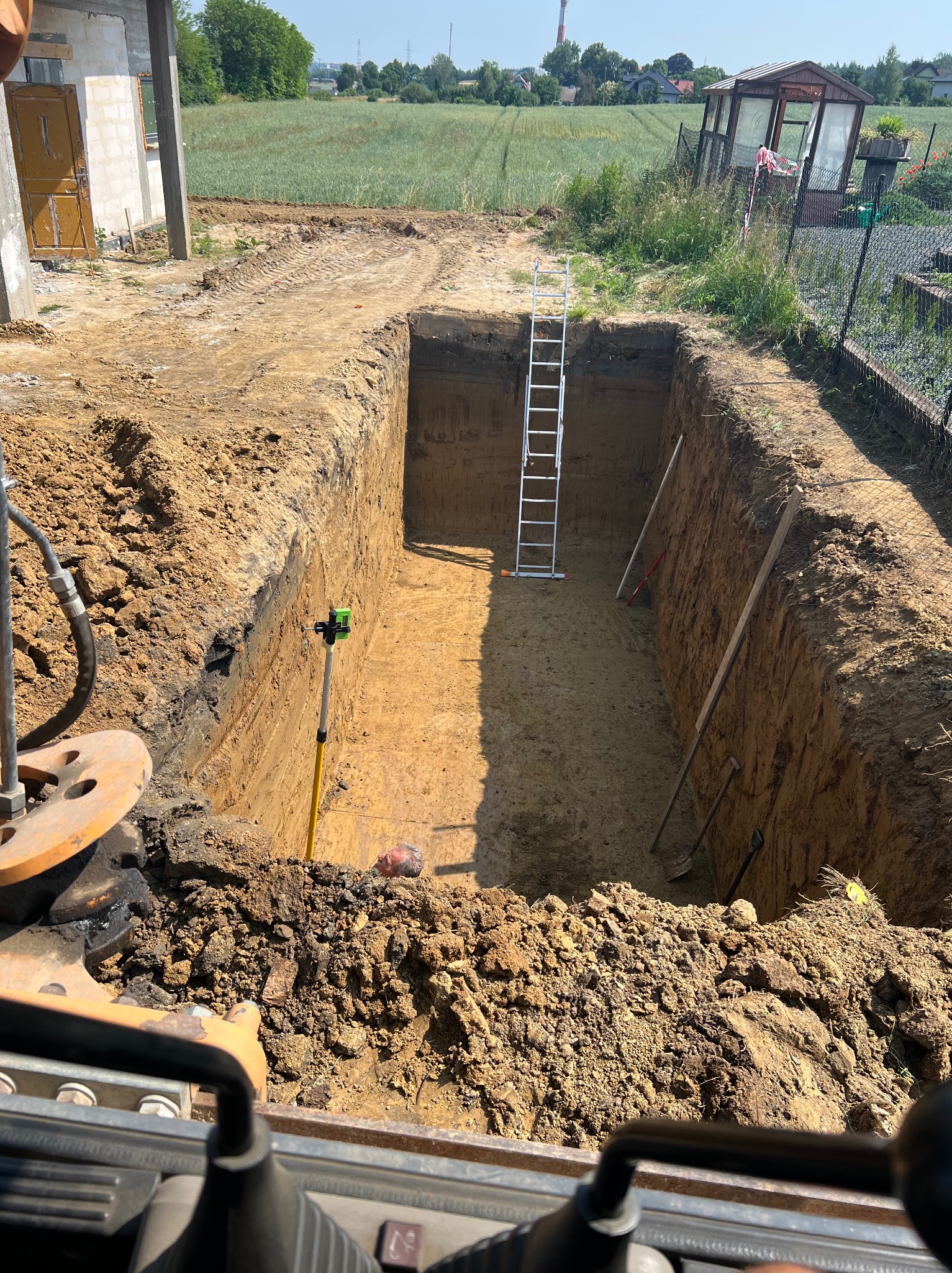 Głęboki wykop w ziemi, widoczna drabina, poziomica laserowa i narzędzia, w tle budynek w trakcie budowy i zielone pole, ujęcie z kabiny maszyny budowlanej.