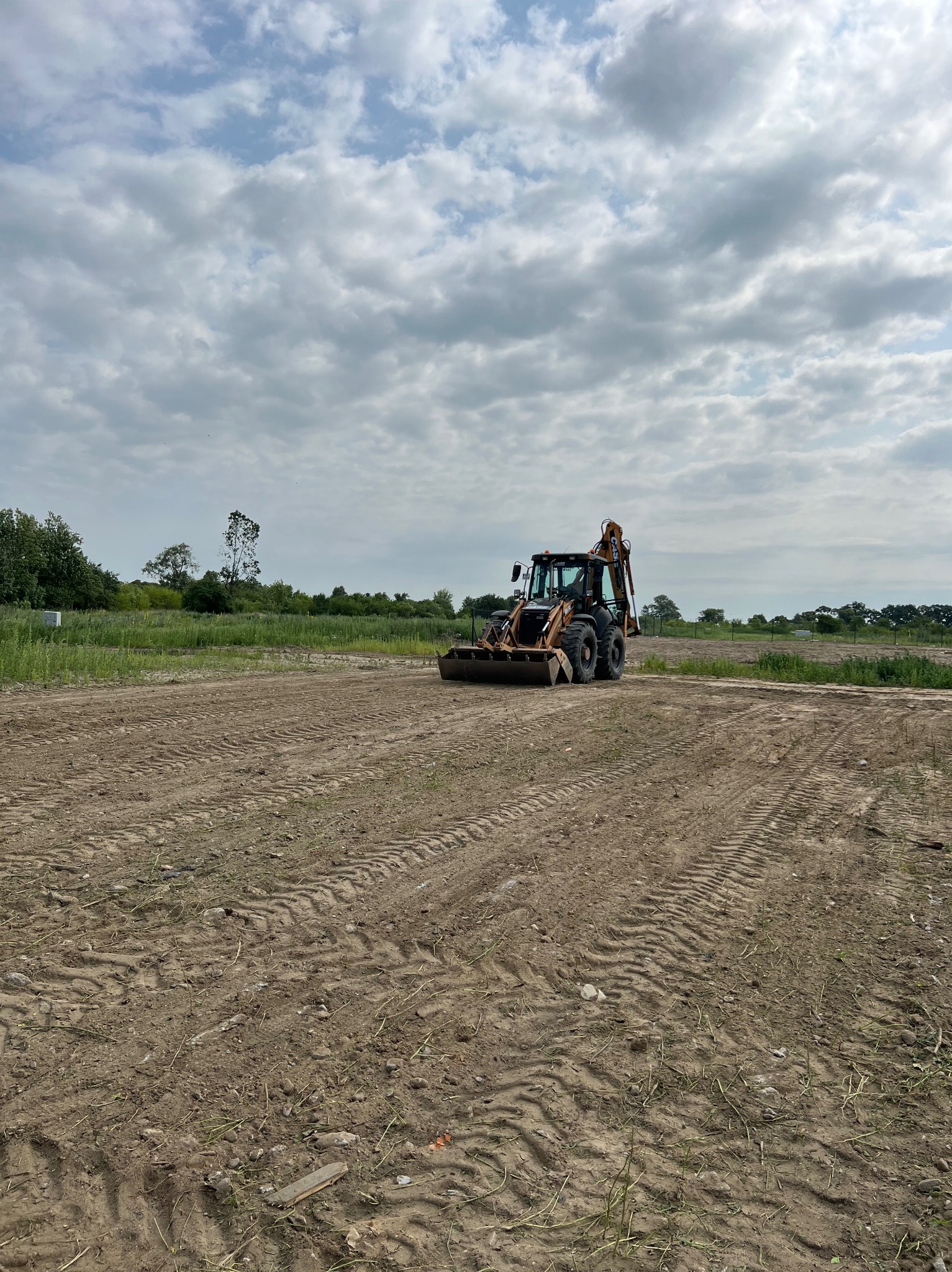 Pomarańczowo-czarna koparko-ładowarka na przygotowanym, brązowym terenie z wyraźnymi śladami opon, pod pochmurnym niebem. Widoczne drzewa w tle.