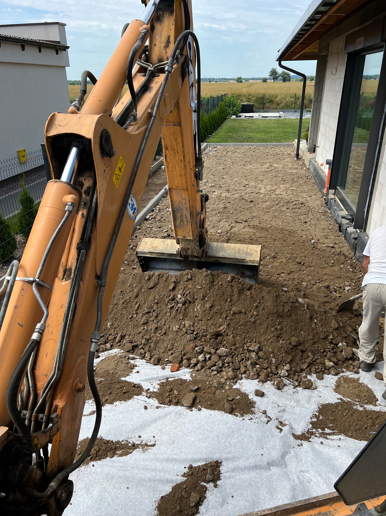 Pomarańczowa koparka CASE nabiera ziemię na białej geowłókninie, widoczny operator z łopatą, przygotowanie terenu pod budowę domu z dużym oknem.