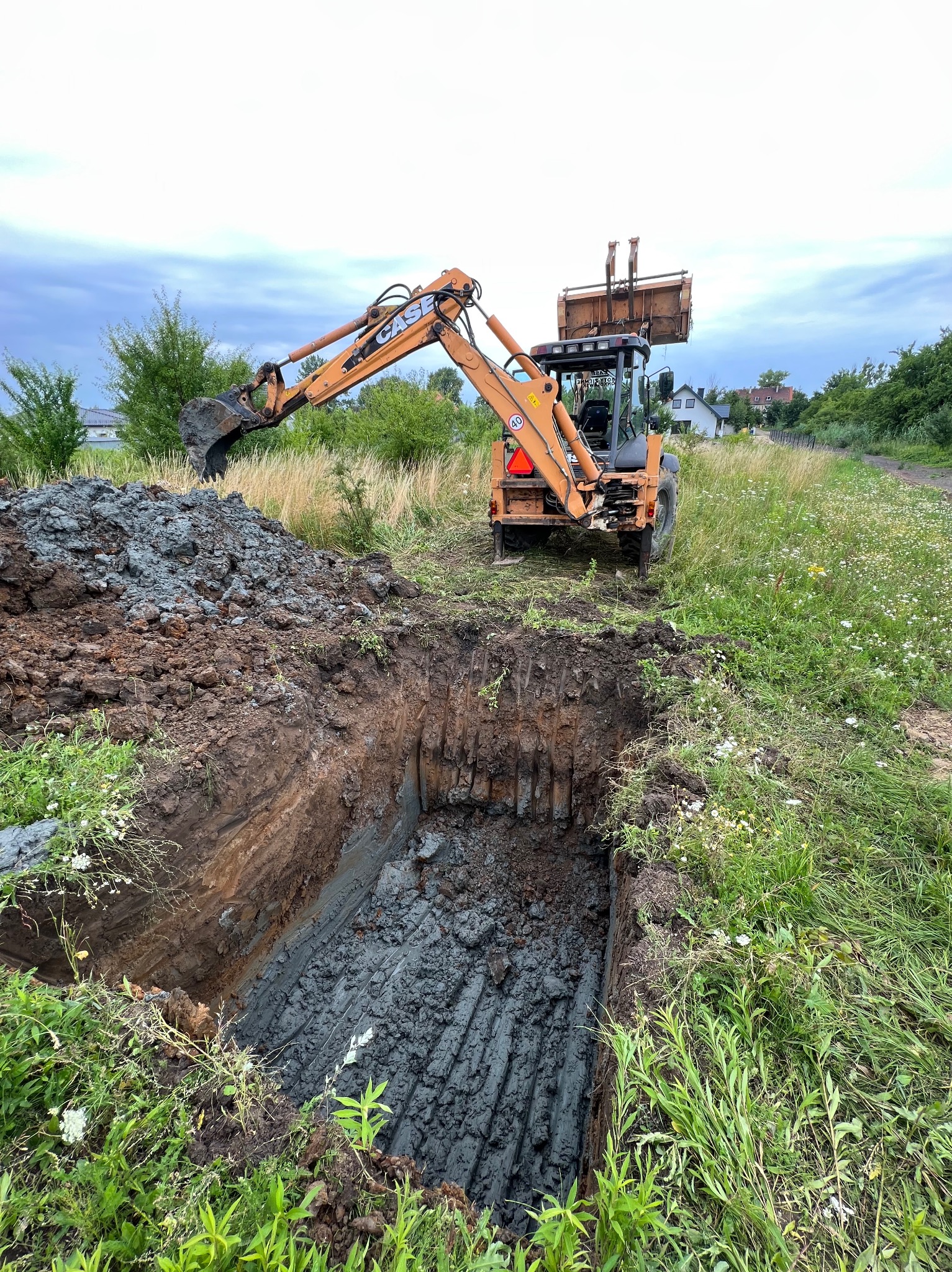 Pomarańczowa koparko-ładowarka Case na tle zieleni, wykopany głęboki dół z widocznymi śladami łyżki koparki na dnie, obok sterta wykopanej ziemi.