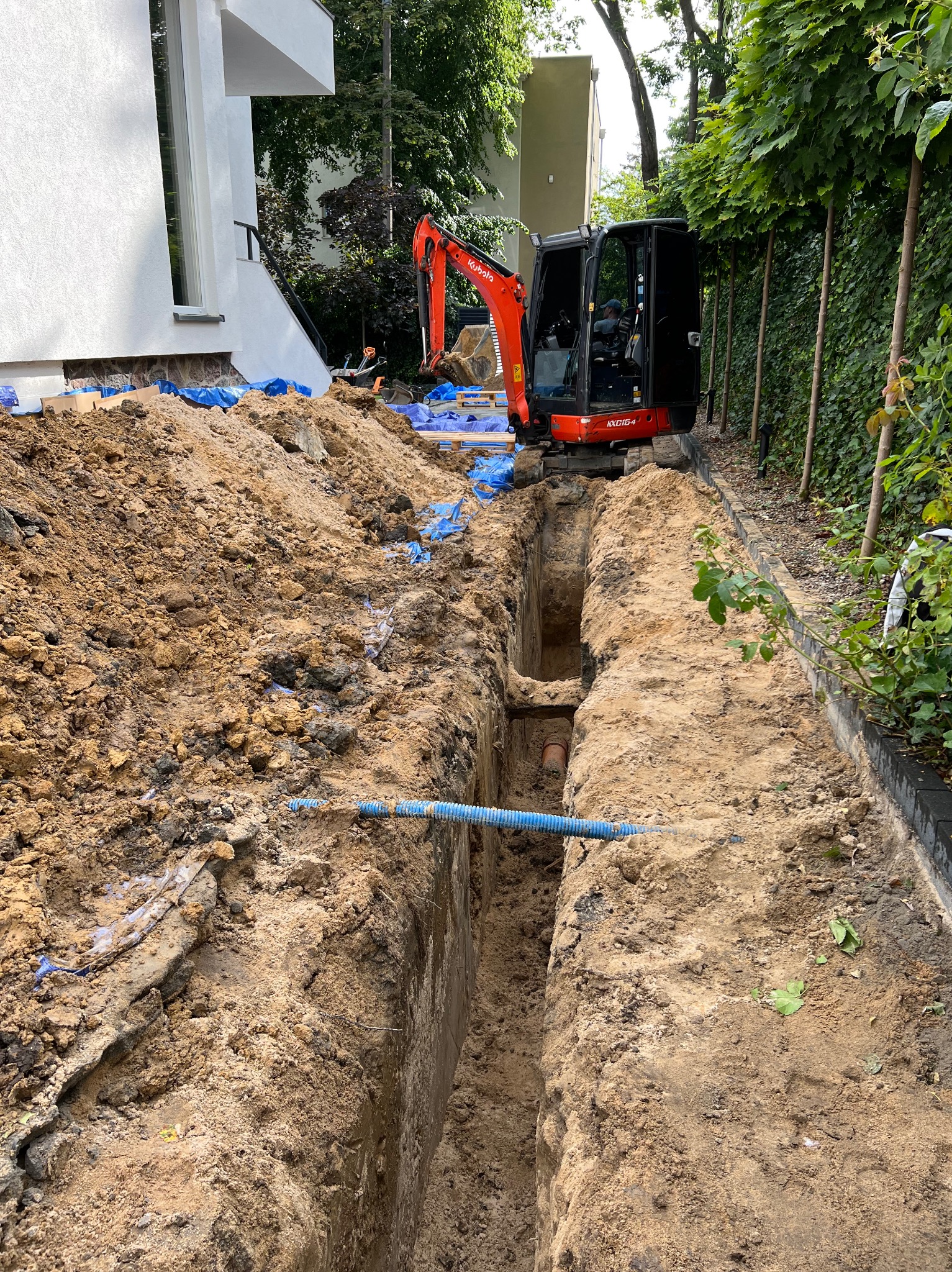 Wąski wykop w ziemi, z widoczną rurą wewnątrz i małą koparką Kubota obok, zabezpieczony niebieską folią, na tle budynku i zieleni.