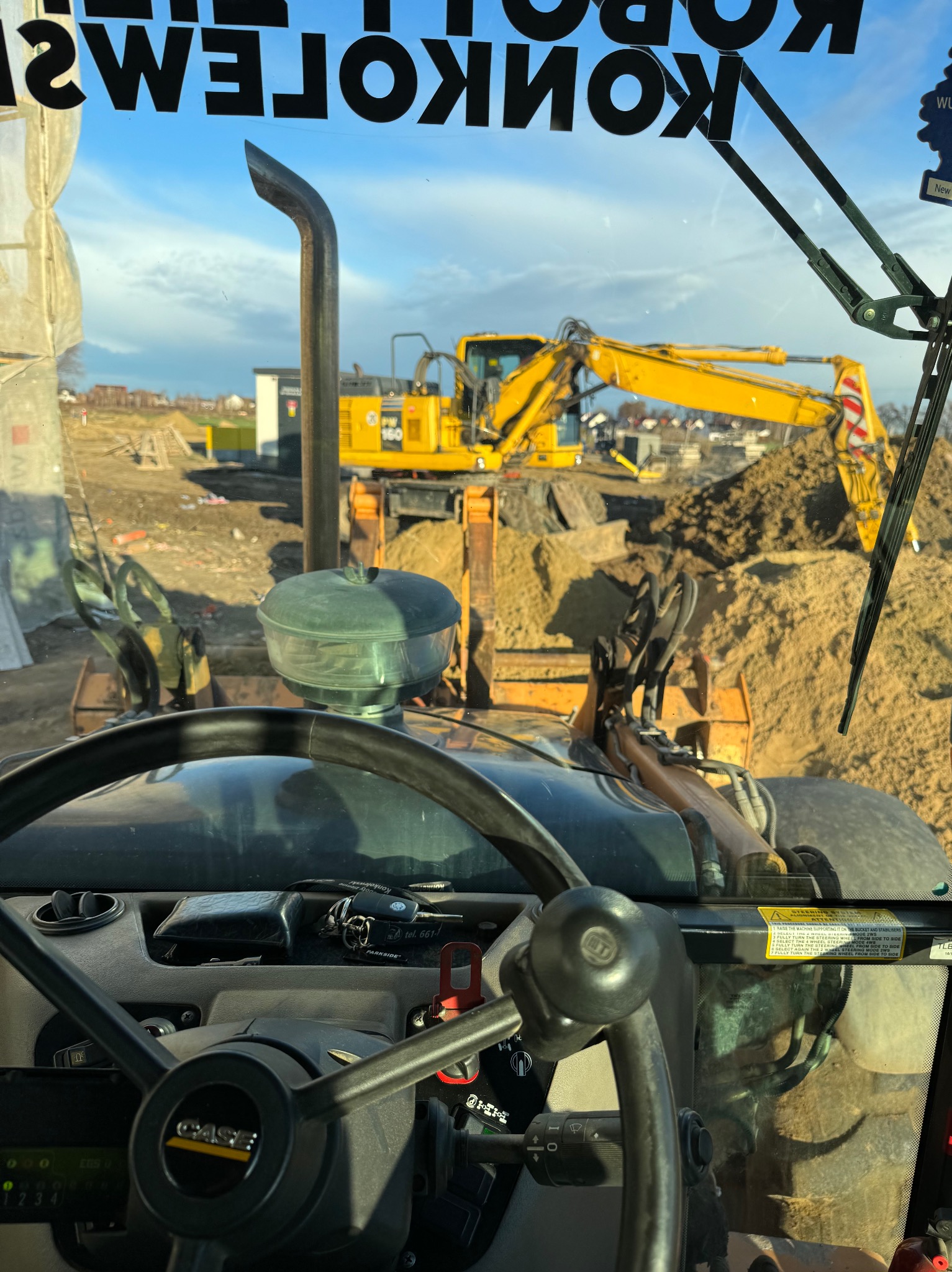 Widok z kabiny operatora na plac budowy z dwoma żółtymi koparkami, jedna z koparek pracuje przy usypanej górze piachu, w tle widać zabudowę w trakcie realizacji.