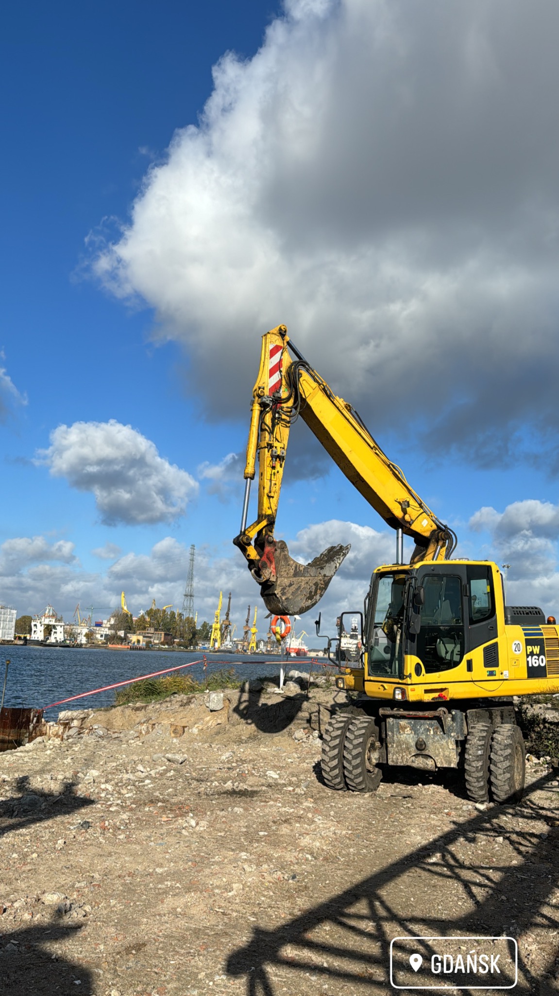 Żółta koparka kołowa Komatsu PW160 z uniesioną łyżką nabierającą ziemię na tle portu w Gdańsku i błękitnego nieba z chmurami.