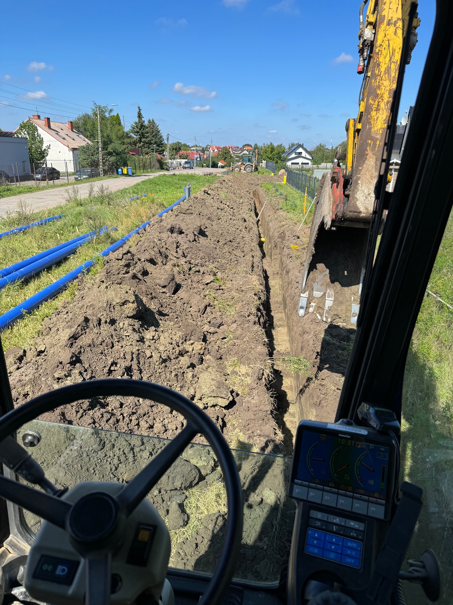 Wykop wąskoprzestrzenny widziany z kabiny koparki, z ułożonymi niebieskimi rurami obok, w tle zabudowa jednorodzinna.