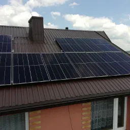 Brązowy dach z blachy trapezowej z zamontowanymi panelami słonecznymi, komin z blachy w kolorze dachu, widok z góry na dom i okoliczne zabudowania w słoneczny dzień.