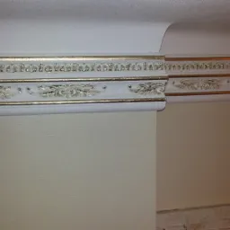 Dekoracyjna listwa przysufitowa z ornamentami w kolorze białym i złotym, zamontowana na styku ściany i sufitu w jasnym pomieszczeniu.