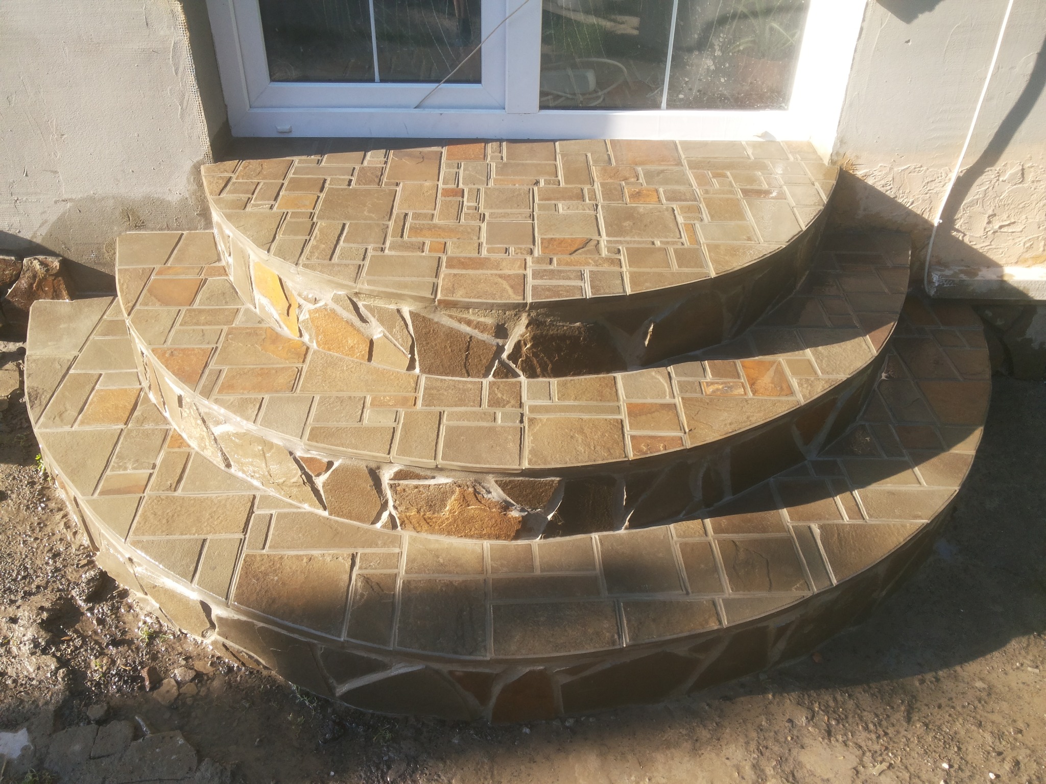 Trzy półokrągłe schody wejściowe wyłożone mozaiką z kamienia naturalnego w odcieniach beżu i brązu, prowadzące do białych drzwi balkonowych.