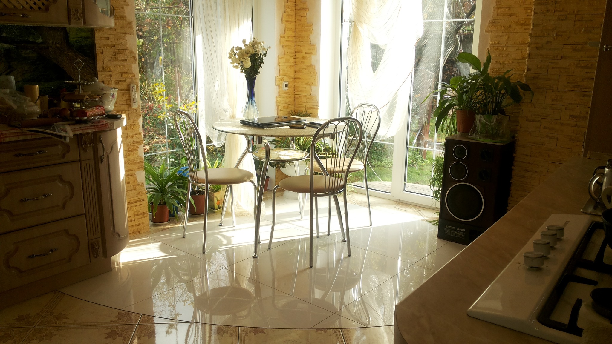 Jasna kuchnia z dużym oknem, okrągłym stołem, metalowymi krzesłami i błyszczącą podłogą w odcieniach beżu i brązu. Na stole leżą laptop i książki, a w tle widać rośliny i dekoracyjną ścianę...