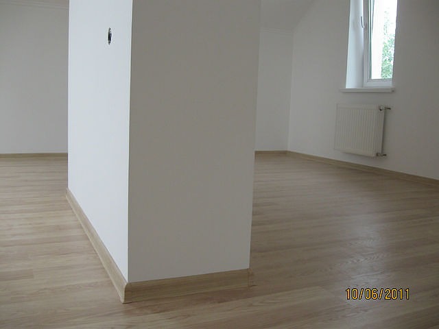 Pomalowane na biało wnętrze z nową podłogą z paneli w odcieniach jasnego drewna, widoczna listwa przypodłogowa i grzejnik pod oknem.