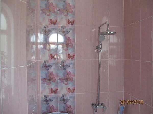 Różowa łazienka z płytkami w motyle, bateria prysznicowa z deszczownicą, widoczne odbicie okna w glazurze.