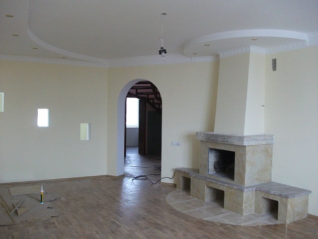 Jasne wnętrze z kominkiem obłożonym beżowym kamieniem, parkietem i ozdobnym sufitem z elementami sztukaterii, widoczne materiały budowlane na podłodze.