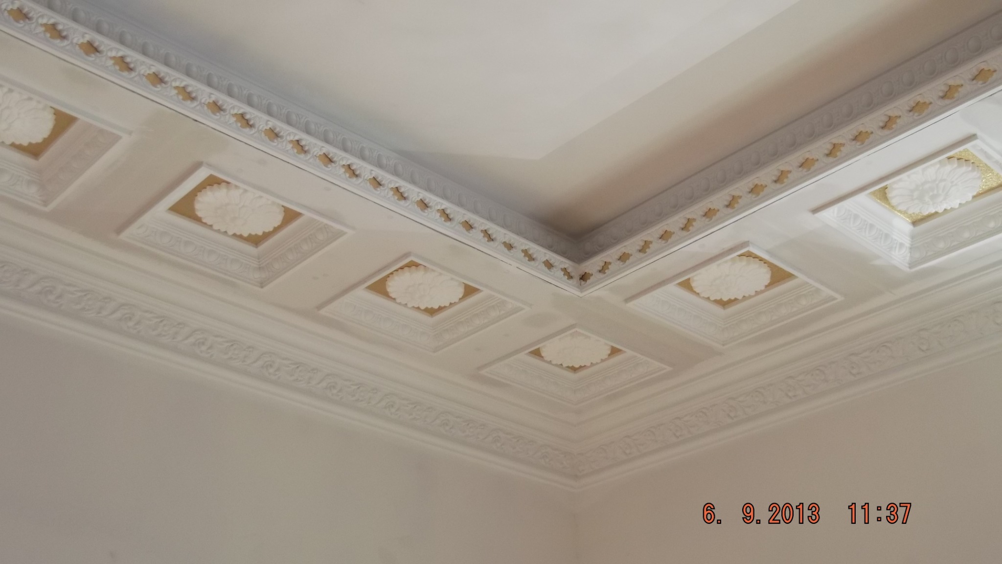 Dekoracyjny sufit z białymi, kwadratowymi kasetonami z motywem kwiatowym i złoconym obramowaniem, wykończony sztukaterią.