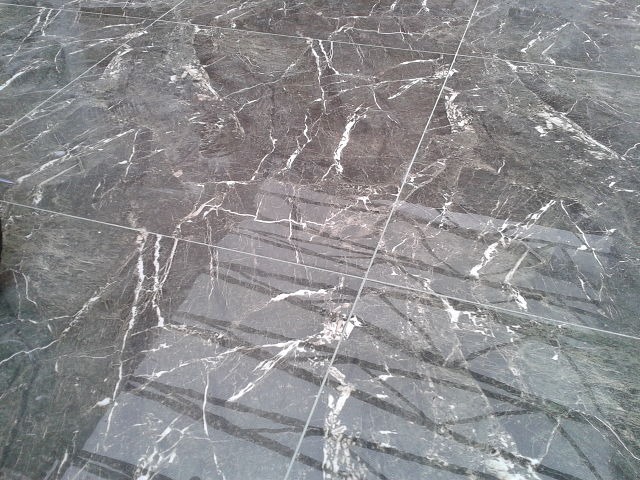 Połyskująca, ciemnoszara marmurowa podłoga z wyraźnym białym użyleniem, ułożona z płytek o prostych krawędziach, widoczne odbicia światła na powierzchni.