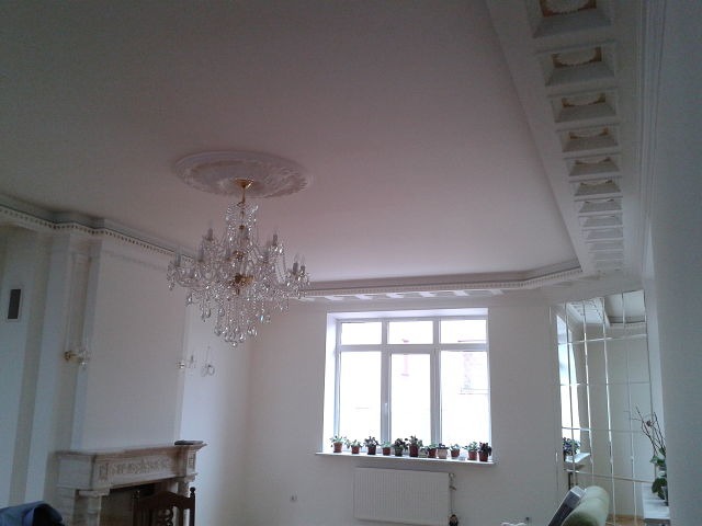 Jasne wnętrze pokoju po remoncie z ozdobnym sufitem, kryształowym żyrandolem, marmurowym kominkiem i lustrami na ścianie.