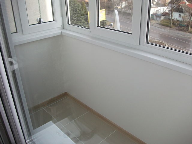 Białe, przeszklone okno balkonowe z widokiem na ulicę, wykończone płytkami podłogowymi i gładkimi, kremowymi ścianami.