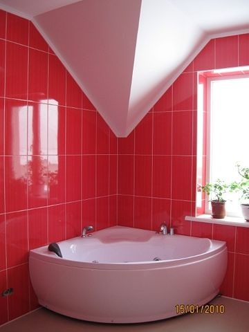Biała narożna wanna z hydromasażem w łazience wyłożonej intensywnie czerwoną, pionowo ułożoną glazurą. Nietypowa konstrukcja sufitu z wnęką w kształcie trójkąta. Na parapecie okna doniczki...