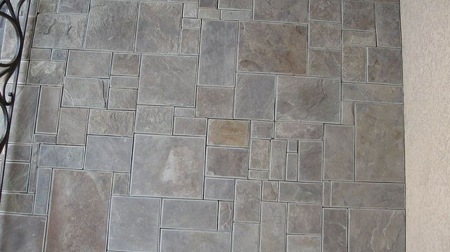 Ściana wyłożona nieregularnymi, szarymi płytkami imitującymi kamień, z fragmentem czarnej, ozdobnej balustrady po lewej stronie i jasnym, teksturowanym tynkiem po prawej.