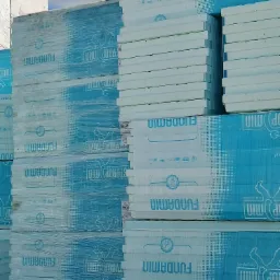 Stosy białych płyt styropianowych Fundamin, częściowo owiniętych w niebieską folię z nadrukiem, składowane na zewnątrz.