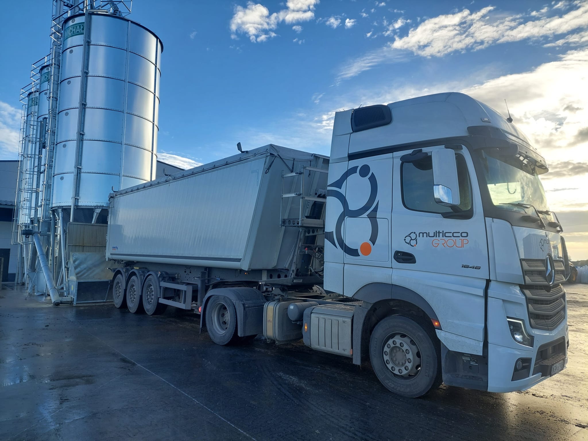 Ciężarówka z naczepą typu wywrotka na tle silosów zbożowych, logo Multicco Group na kabinie.