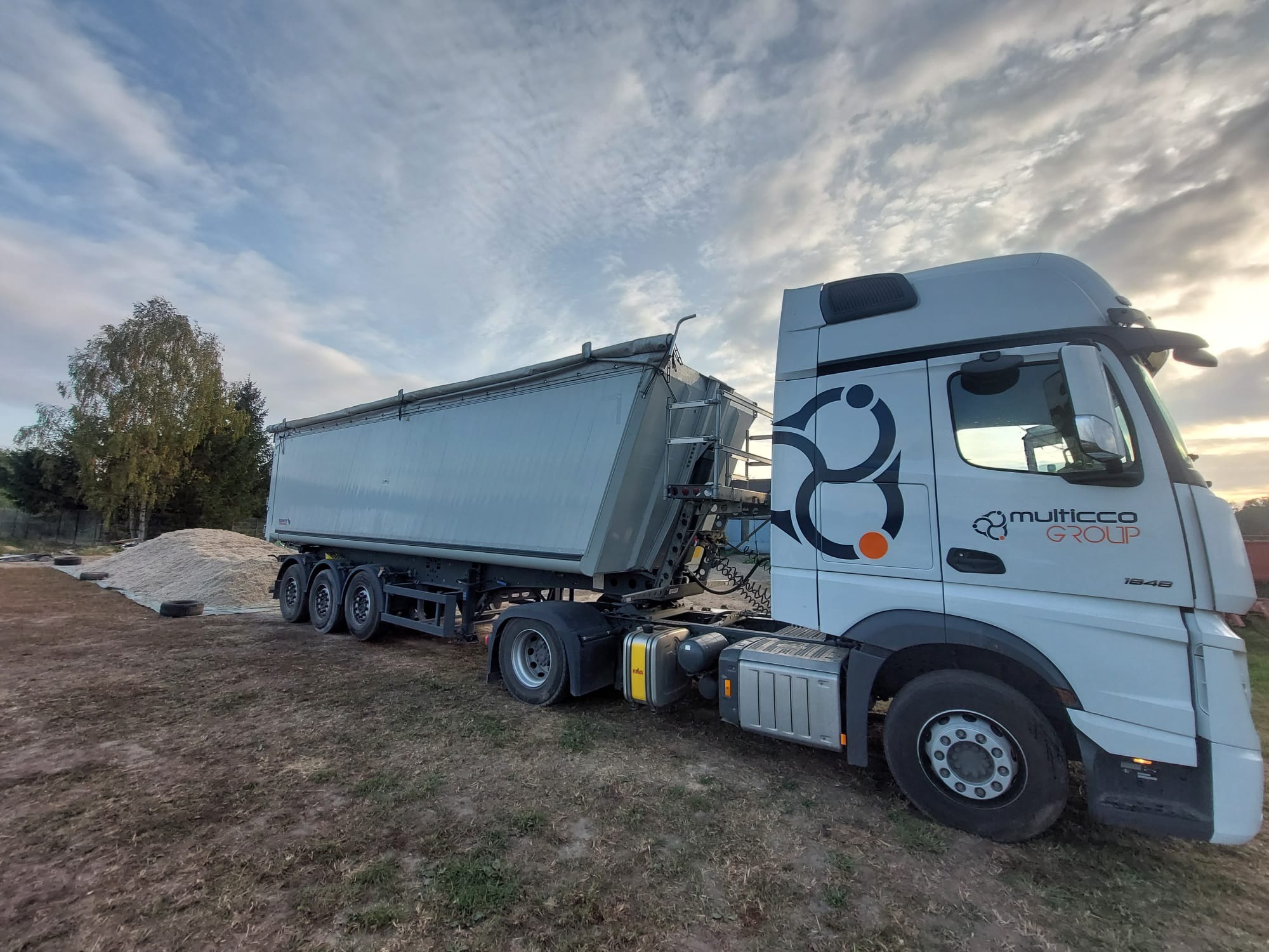 Ciężarówka z naczepą typu wywrotka, zaparkowana na ziemi obok pryzmy piasku zabezpieczonej oponami, widoczny branding 'multicco GROUP' na kabinie
